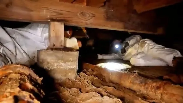 Worker inspecting rusted cast-iron pipe under a house